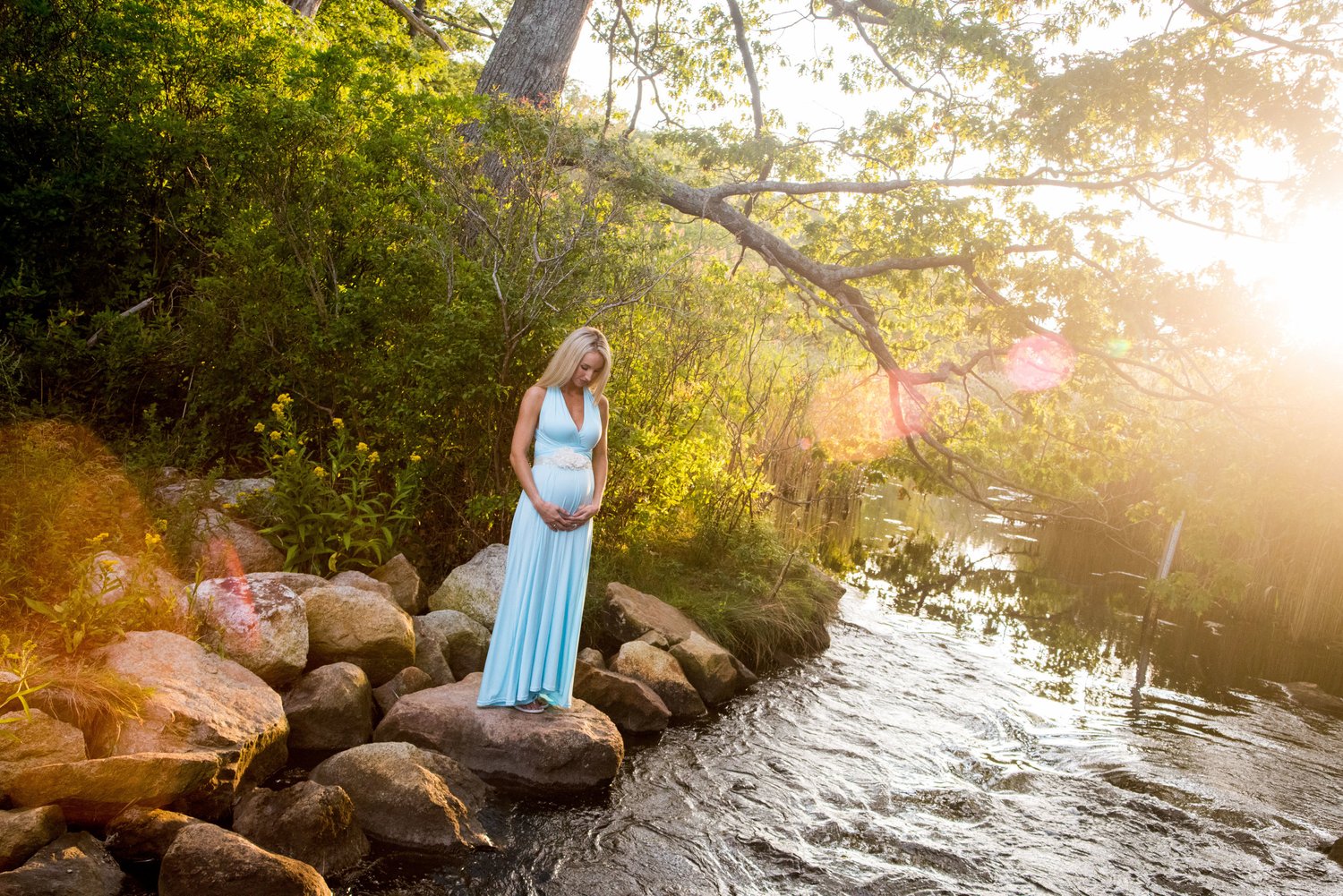 Pregnant woman in a pale blue gown stands on riverside rocks in sun flare