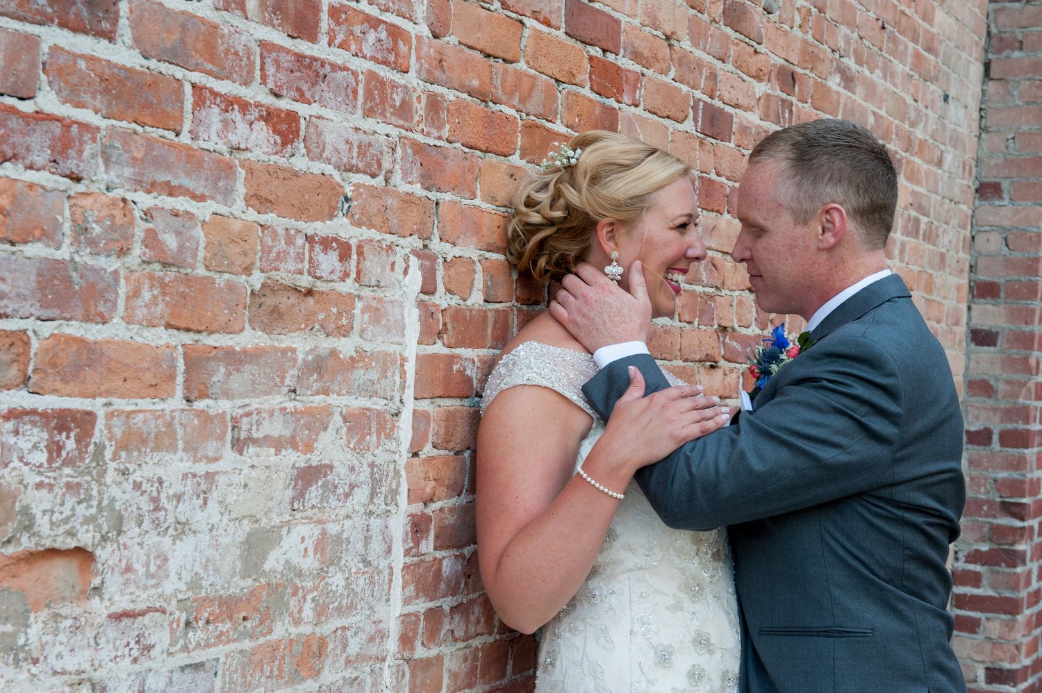 Bride laughs against a weathered brick wall as the groom leans in